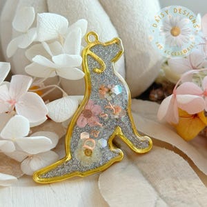Può includere: Un ciondolo a forma di cane con una cornice in metallo dorato, riempito di glitter argento e decorato con piccoli fiori pressati. Il nome Daisy è scritto in lettere color rame al centro. È presente un piccolo occhiello circolare sulla parte superiore.
