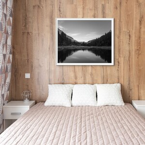 May include: A bedroom with a wooden wall, a bed with white pillows and a beige quilted bedspread, and a black and white photograph of a lake and mountains hanging on the wall.