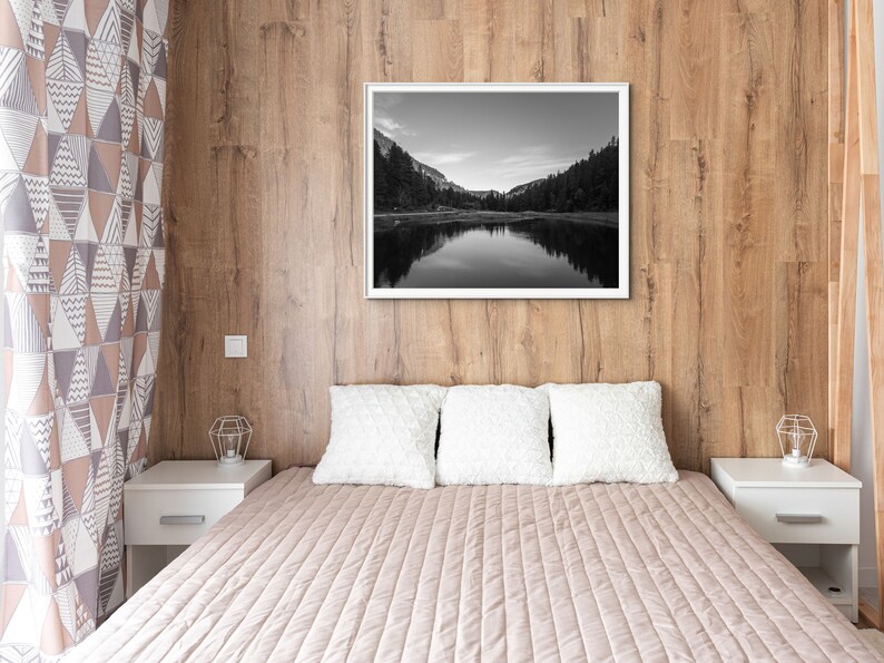 May include: A bedroom with a wooden wall, a bed with white pillows and a beige quilted bedspread, and a black and white photograph of a lake and mountains hanging on the wall.