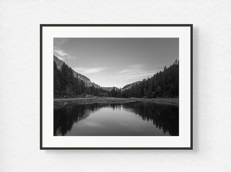 May include: A black and white photograph of a lake reflecting the surrounding trees and mountains.