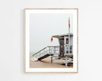Peut inclure: Impression encadrée d'un poste de sauveteur sur une plage de sable. La structure en bois bleu clair est dotée d'un escalier, d'une petite fenêtre et d'un drapeau américain. Une bouée de sauvetage orange est suspendue au toit. Une planche de surf repose sur le sable.