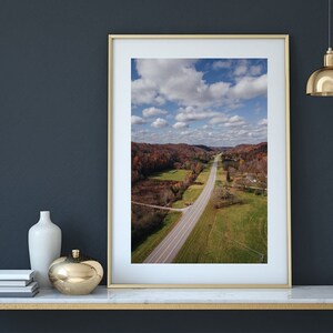 May include: An aerial photograph of a long, straight road winding through a valley with green fields and trees. The sky is blue with white clouds.