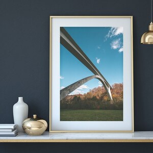 May include: A framed photograph of a concrete bridge with two arches against a blue sky with white clouds. The bridge is surrounded by green grass and trees with fall foliage.