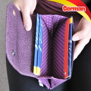 May include: A purple fabric wallet with a snap closure, featuring a patterned interior lining with multiple vertical slots. The wallet is open, revealing several blue and red cards inserted into the compartments, showcasing the internal structure.