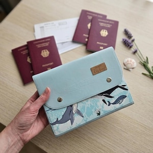 Könnte beinhalten: Ein hellblauer Dokumentenorganizer mit Wal-Motiv, einer Klappe mit zwei Metalldruckknöpfen und einem rechteckigen braunen Etikett mit der Aufschrift "Makers Day Design". Der Organizer ist zusammen mit vier bordeauxroten Reisepässen abgebildet.