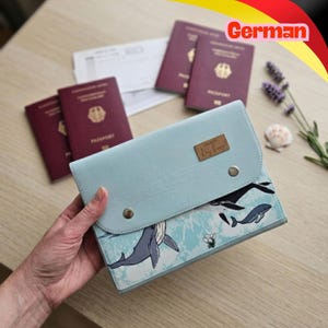 Puede incluir: Un portapasaportes azul claro con estampado de ballenas y una etiqueta rectangular marrón con el texto "Day Design". El estuche tiene una solapa con dos botones de presión de metal plateado. Al fondo hay tres pasaportes color burdeos.