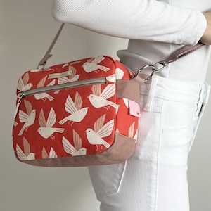May include: A rectangular crossbody bag featuring a vibrant orange fabric with a repeating pattern of white and light pink stylised birds. The bag includes a front zippered pocket, a brown base, and a matching brown adjustable shoulder strap.