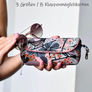 Könnte beinhalten: Ein rechteckiges Brillenetui aus Stoff mit einem Blumen- und Beerenmuster in Blau-, Rosa- und Schwarztönen sowie einem rosa Rand. Das Etui hat eine seitliche Schlaufe mit einem Metallclip. Der Text oben lautet 3 Größen / 8 Klappenmöglichkeiten.
