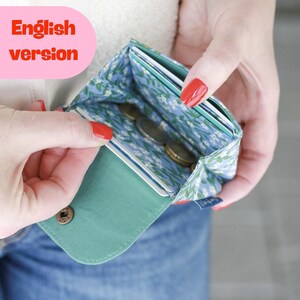 May include: A small fabric wallet featuring a light blue interior lining with a white and green floral pattern. The wallet includes multiple slots for cards and coins, a solid teal green flap, and a small metallic snap button closure.