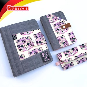 May include: A set of three items including a large notebook, a smaller notebook, and a wallet, all featuring a cream fabric with a pink and purple cat face pattern and dark grey plaid fabric accents. The large notebook has a black label with the text BORN TO BE AWESOME.