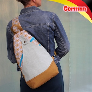 May include: A sling backpack featuring a patterned fabric with gold and white circular motifs, a solid light grey central panel, and a solid brown base. The bag includes bright blue zipper accents, a brown strap, and a metal snap button closure.