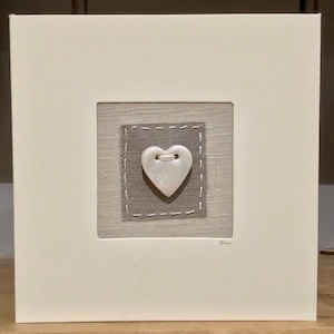 May include: A white card with a grey fabric square in the centre. The fabric square has a white ceramic heart with a small hole in the top. The card is signed "Sue".