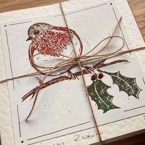 May include: A stack of cream-colored cards tied with twine. Each card features a brown and red illustration of a robin perched on a branch with holly. The cards are bundled together with a twine bow.