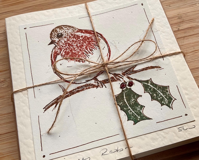 May include: A stack of cream-colored cards tied with twine. Each card features a brown and red illustration of a robin perched on a branch with holly. The cards are bundled together with a twine bow.