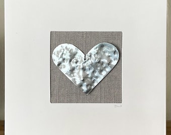 May include: A silver heart-shaped metal cutout is displayed on a beige linen background. The heart is textured and has a hammered finish. The heart is framed in a white mat and a white frame.
