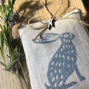 May include: A linen sachet featuring a blue hare print, adorned with twine and a wooden star. Fresh lavender sprigs are arranged on a wooden surface. The sachet is filled with dried lavender, creating a rustic, natural aesthetic.