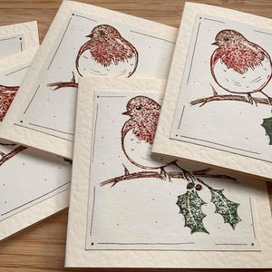 May include: A collection of square greeting cards with a hand-drawn illustration of a robin perched on a branch with holly. The robin has a red breast and is set against a cream-colored background with a decorative border.