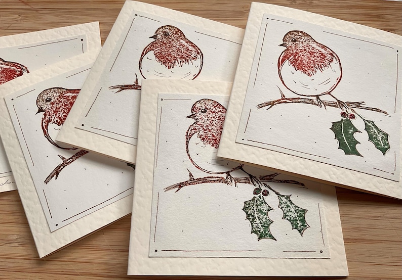 May include: A collection of square greeting cards with a hand-drawn illustration of a robin perched on a branch with holly. The robin has a red breast and is set against a cream-colored background with a decorative border.