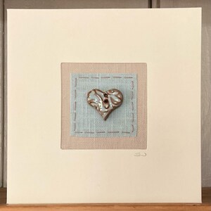 May include: A framed artwork with a heart-shaped button. The button is glossy brown with blue details, centered on a pale blue square stitched onto a beige fabric. The artwork is set against a cream background.