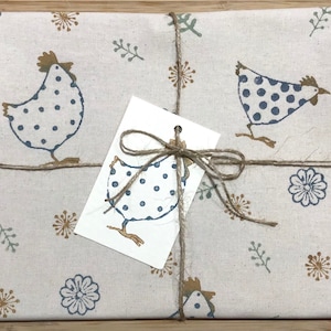 May include: A beige linen tea towel with a brown twine bow. The towel features a repeating pattern of blue and white polka dot chickens and brown floral designs.