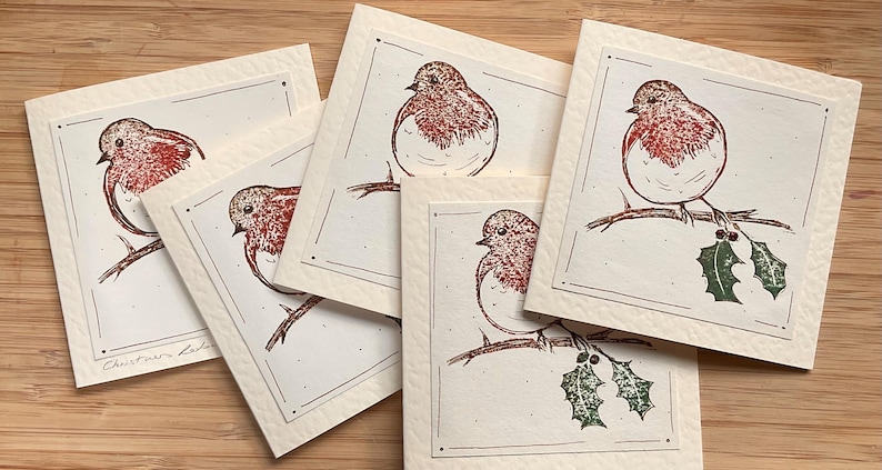 May include: A collection of cream-colored square cards featuring a detailed illustration of a robin bird perched on a branch. The bird has a red breast and brown details. Some cards include green holly. The text "Christmas Cards" is visible on one card.