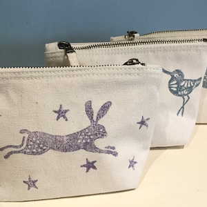 May include: Three white canvas zippered pouches with different animal designs. The pouch on the left has a purple hare with stars. The middle pouch has a blue bird with stars. The pouch on the right has a purple hare with stars.