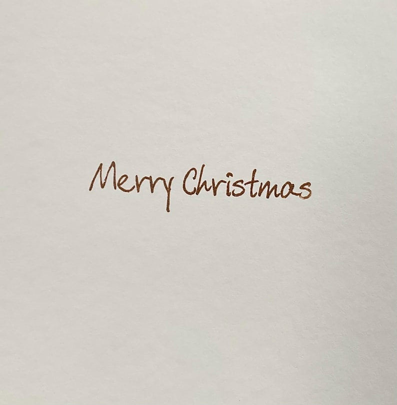 May include: A white card with the handwritten phrase "Merry Christmas" in a brown script. The text is centered and takes up most of the frame, with a simple and elegant design.