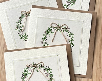 May include: A stack of handmade greeting cards featuring a watercolor wreath design. Each card has a cream-colored textured paper with a green border and a wreath of green leaves, berries, and a twine bow. The cards are paired with brown envelopes.