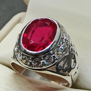 May include: A silver ring with a large, oval-shaped, red gemstone surrounded by smaller, clear gemstones. The ring has an intricate design on the band.