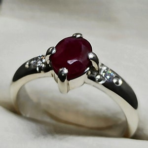 May include: A silver ring featuring a red oval ruby gemstone and two smaller clear gemstones set into the band.