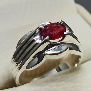 Puede incluir: Un anillo de plata con una piedra preciosa roja engastada en garras. El anillo tiene una banda texturizada.