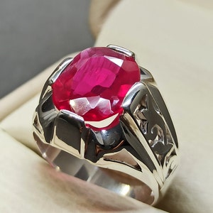 Könnte beinhalten: Ein silberner Ring mit einem großen, ovalen, roten Edelstein, der in der Mitte eingesetzt ist. Der Ring hat komplizierte, detaillierte Designs auf der Band.