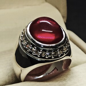 May include: A silver ring with a large, oval-shaped, red gemstone set in a bezel setting. The ring is decorated with a band of smaller, clear gemstones.