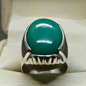 May include: A silver ring featuring a large, oval-shaped green gemstone set in the centre. The ring has a detailed, textured band.