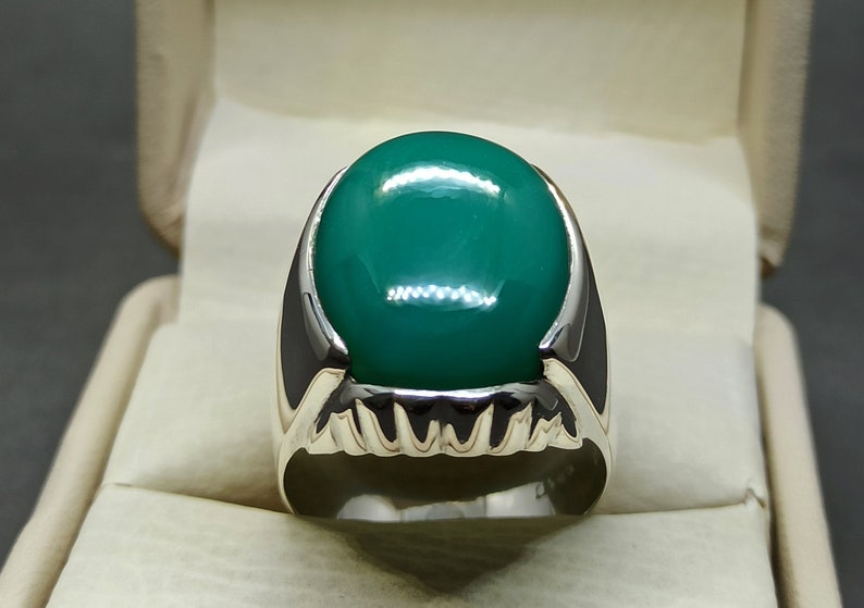 May include: A silver ring featuring a large, oval-shaped green gemstone set in the centre. The ring has a detailed, textured band.