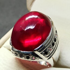 May include: A silver ring with a large, oval-shaped, red gemstone set in the center. The band is decorated with smaller, black gemstones.