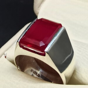 May include: A silver ring featuring a large, rectangular, faceted, deep red gemstone. The ring has a thick band and a modern, geometric design. The gemstone is the focal point, creating a bold statement.