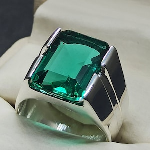 Puede incluir: Un anillo de plata con una gran piedra preciosa verde esmeralda. El anillo tiene una banda gruesa con un acabado liso y pulido.