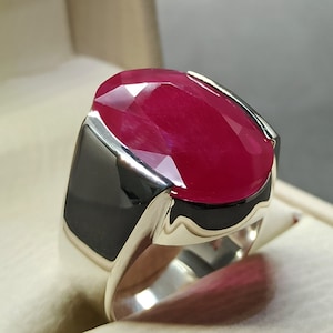 Può includere: Anello con un grande rubino ovale di colore rosso intenso. La pietra è incastonata in una fascia argentata con dettagli neri. L'anello è presentato su uno sfondo neutro, evidenziando la brillantezza del rubino.