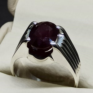 May include: A silver ring with a dark red oval gemstone set in a prong setting. The ring has a textured band with a series of vertical lines.