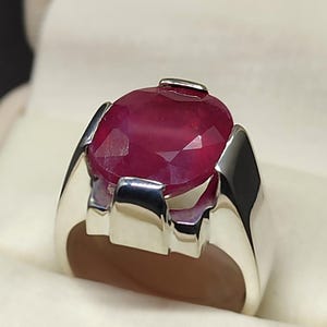 May include: A silver ring with a large, oval-shaped ruby gemstone. The ruby is a deep red color and has a faceted cut.