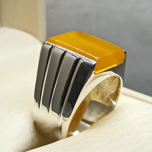 May include: A silver ring with a yellow gemstone set in a square shape. The ring has a textured band with three vertical lines.