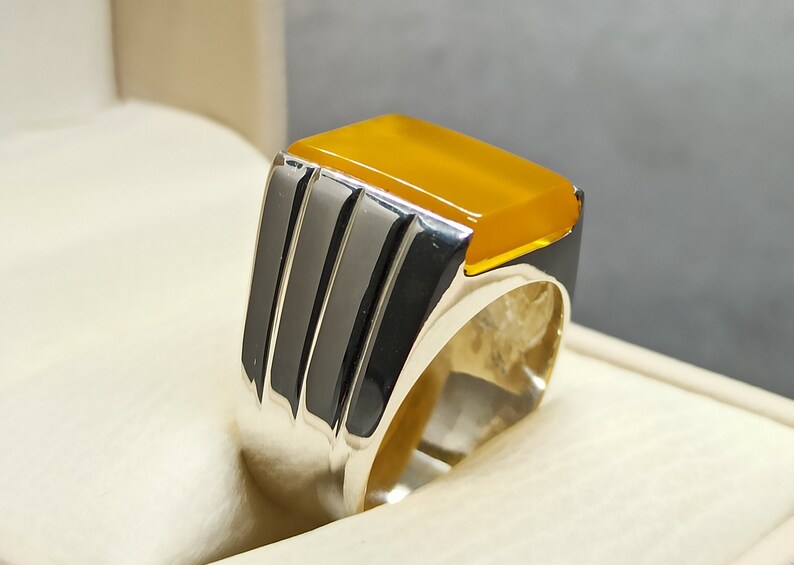 May include: A silver ring with a yellow gemstone set in a square shape. The ring has a textured band with three vertical lines.