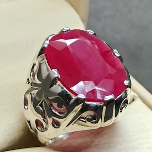 May include: A silver ring featuring an oval-shaped, faceted, red gemstone set in an ornate, openwork band.
