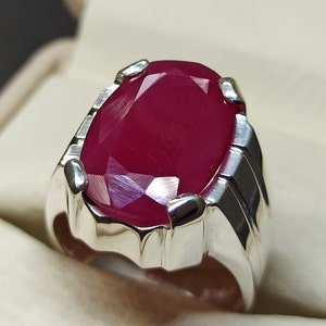 May include: A silver ring featuring a large, oval-shaped ruby gemstone. The ruby is a deep red colour and has a faceted cut.