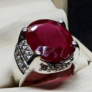 Könnte beinhalten: Ein silberner Ring mit einem großen, ovalen Rubin-Edelstein, der in der Mitte sitzt. Der Ring ist mit kleineren, weißen Edelsteinen um den Rubin herum verziert.