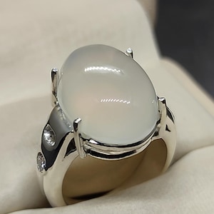 Könnte beinhalten: Ein silberner Ring mit einem großen, ovalen, weißen Edelstein. Der Ring hat ein detailliertes Design mit kleinen Akzenten.