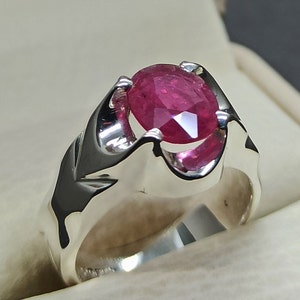 May include: A silver ring with a large, oval-shaped, pink ruby gemstone set in the center. The ring has a detailed, textured band.