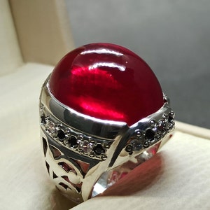 May include: A silver ring with a large, round, red gemstone set in the center. The ring is decorated with smaller white and black gemstones around the band.