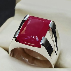 May include: A silver ring with a large, rectangular, deep red gemstone. The ring has a polished, modern design with a thick band and angular accents around the stone. The ring is displayed in a white jewelry box.